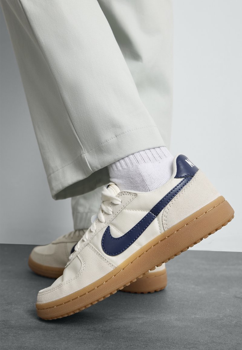 Baskets beiges avec un Swoosh Nike bleu marine et des semelles en gomme, portées avec des chaussettes blanches et un pantalon gris clair sur un sol gris.