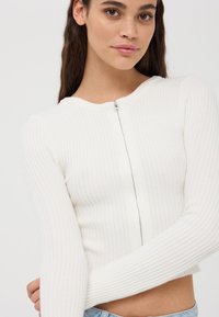 Biały prążkowany sweter na zamek błyskawiczny, krótki, z okrągłym dekoltem i długimi rękawami, o gładkiej fakturze i subtelnym połysku.