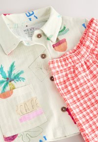 Short-sleeve button-up shirt in light beige with colorful plant designs and a chest pocket, paired with red and white checkered shorts.