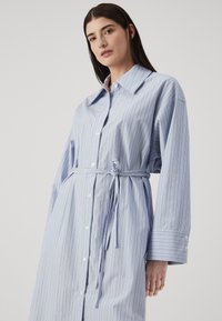 Vestito camicia blu chiaro con un motivo a righe orizzontali e verticali, vita con lacci e maniche lunghe con ampi polsini.