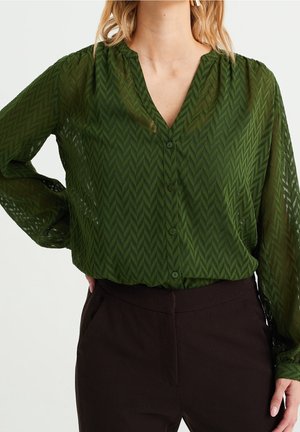 Femme portant un chemisier transparent vert foncé à motif chevrons avec boutons et un pantalon noir taille haute sur fond blanc.