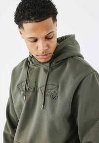 Haut à capuche vert olive avec un cordon de serrage et un logo brodé "Perfect Paris". Il présente une coupe décontractée et un tissu doux et texturé.