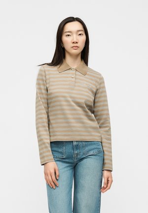 Femme aux longs cheveux foncés portant un polo rayé beige et bleu clair à manches longues et un jean bleu taille haute, debout devant un fond blanc.