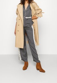 Trench coat beige sopra una tuta nera a fantasia con strisce bianche ai lati. Stivali alla caviglia marroni completano l'outfit.