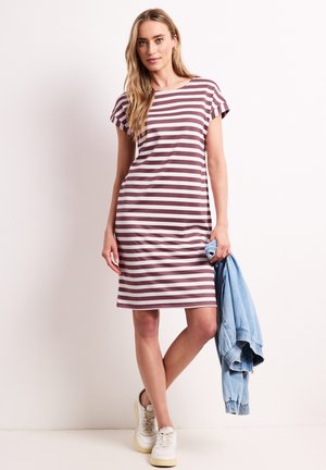 Mujer con un vestido de manga corta hasta la rodilla, con rayas horizontales en granate y blanco, zapatillas blancas, sosteniendo una chaqueta vaquera azul claro.