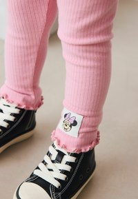Child wearing pink ribbed leggings with a Minnie Mouse patch paired with black and white high-top sneakers indoors.