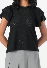 Blusa nera con maniche corte a volant, caratterizzata da un motivo a zigzag di tagli. Abbinata a una gonna plissettata grigia chiara.
