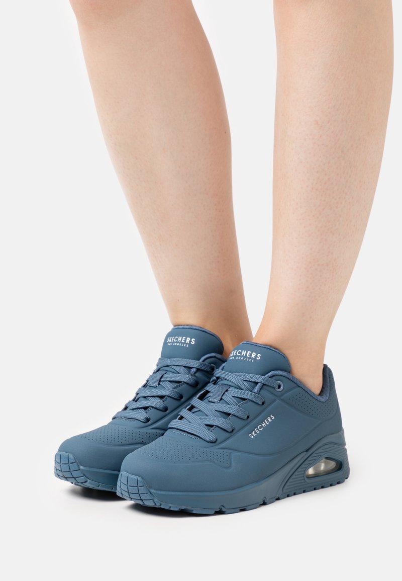 Baskets de sport bleues Skechers portées pieds nus, avec des lacets, des semelles rembourrées et un coussin d'air visible au talon.