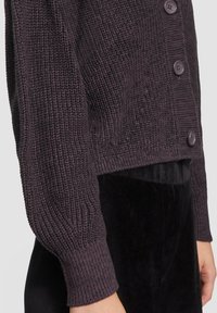 Dunkellilafarbene Strickjacke mit strukturiertem Muster, sechs Knöpfen und gerippten Bündchen, kombiniert mit einer schwarzen Cordhose.