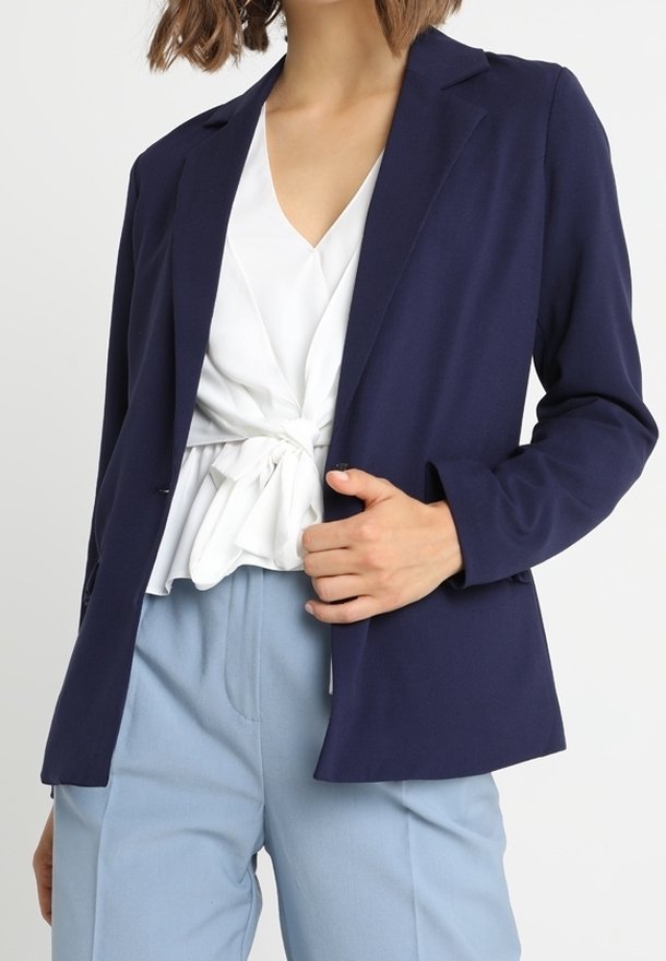 Femme portant un blazer bleu marine sur un chemisier blanc noué et un pantalon bleu clair, une main ajustant le devant du blazer.