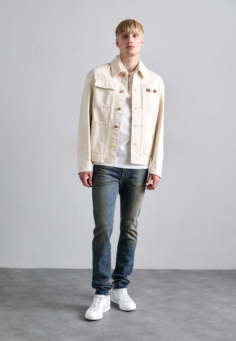 Ljusbeige denimjacka med gyllene knappar, två framfickor, tillsammans med blekta blåjeans och vita sneakers. En enkel vit t-shirt under.