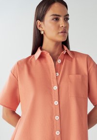 Korallfärgad skjorta med korta ärmar och knappstängning framtill, med vita knappar och en bröstficka. Slät tygtextur, klassisk design.