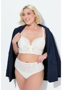Witte kanten bralette met golvende randen en een sluiting aan de voorkant. Hoogzittende bijpassende slip met kanten accenten. Model gehuld in een marineblauwe blazer.