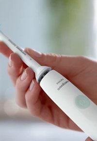 Spazzolino elettrico Sonicare con corpo bianco, pulsante di accensione azzurro chiaro e testa dello spazzolino grigia, tenuto in una mano contro uno sfondo sfocato.