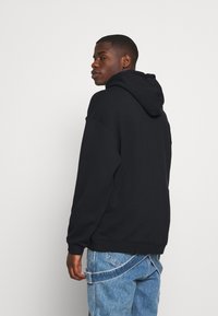 Μαύρη κουκούλα φούτερ με χαλαρή εφαρμογή, διαθέτοντας μαλακή υφή και ριμπ μανσέτες, συνδυασμένη με ανοιχτό μπλε τζιν παντελόνι.