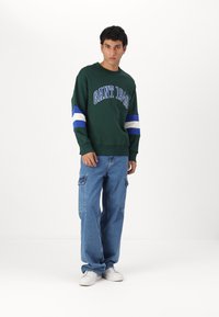 Donkergroen sweatshirt met blauw-witte gestreepte mouwen, voorzien van "GANT 1949" in vette letters. Gecombineerd met lichtgewassen cargobroeken en witte sneakers.