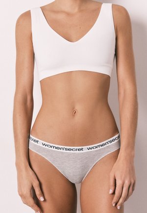 Femme portant un crop top blanc à col en V et un shorty de bikini rayé avec une ceinture à la marque "women’secret", se tenant devant un fond uni.