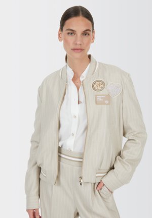 Woman in beige pinstripe jacket and pants with embroidered patches, wearing white button-up shirt, standing with one hand in pocket.