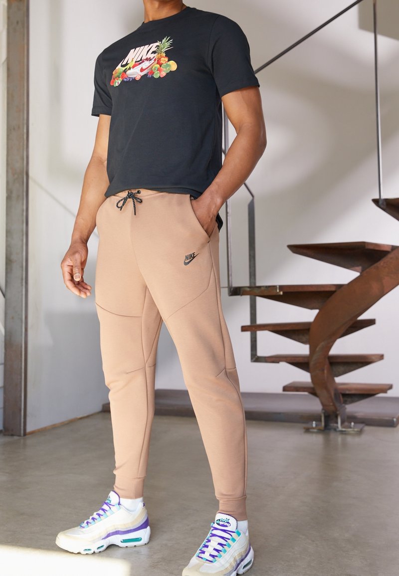 Svart t-shirt med blommig Nike-logotyp, beige joggers med dragsko och fickor, samt flerfärgade Nike Air Max sneakers.