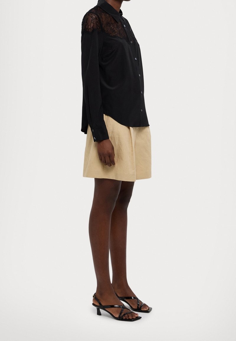 Femme portant un chemisier à manches longues avec un empiècement en dentelle noire, une jupe beige longueur genou, et des sandales à talons noires à brides, debout de profil.