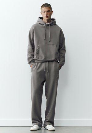 Un mannequin homme se tient debout, les mains dans les poches. Il porte un sweat à capuche gris, un pantalon ample assorti et des baskets blanches, sur un fond de studio uni.