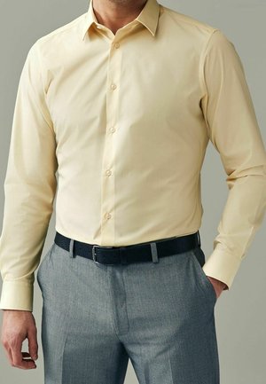 Mann trägt ein blassgelbes langärmliges Hemd, das in graue Hose mit schwarzem Gürtel gesteckt ist, eine Hand in der Tasche.