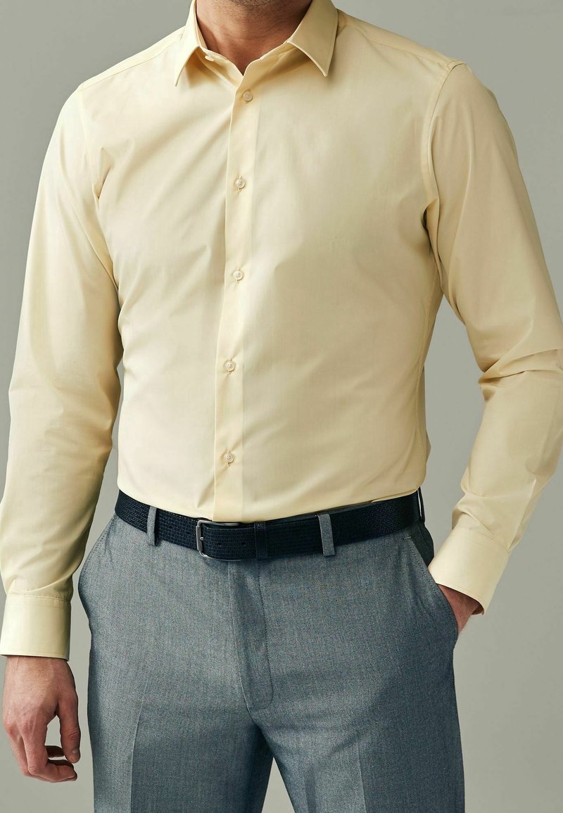 Homme portant une chemise jaune pâle à manches longues rentrée dans un pantalon gris avec une ceinture noire, une main dans la poche.