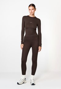 Barna, hosszú ujjú, testhezálló felső és hozzá illő leggings, sima anyagból készült. Figyelemfelkeltő logó a mellkason, sportcipővel és látható fehér zoknikkal párosítva.