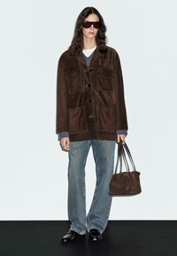 Persona che indossa una giacca di camoscio oversize marrone scuro, jeans azzurri chiari, scarpe nere, grandi occhiali da sole scuri, mentre tiene una borsa a mano in camoscio marrone coordianto.