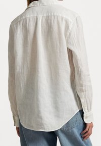 Bílá lněná košile s dlouhými rukávy, špičatým límcem a zakřiveným lemem. Obsahuje manžety na knoflíky a texturovaný povrch.