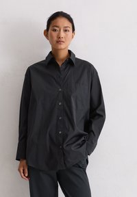 Jonge vrouw met donker haar dat vastgebonden is, gekleed in een losse zwarte blouse met knopen en zwarte broek, tegen een effen witte achtergrond.