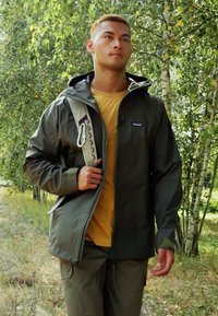 Veste imperméable vert olive avec fermeture éclair à l'avant, capuche ajustable et poches latérales, portée sur une chemise jaune et un pantalon assorti, dans une zone boisée.