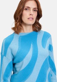 Helle blaue Strickjacke aus einem flauschigen Material mit kräftigen, wellenförmigen Mustern in Dunkelblau. Hat einen runden Ausschnitt und lässig geschnittene Ärmel.