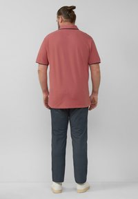 Mann mit zurückgebundenem Haar, der ein staubiges rosa Poloshirt mit kurzen Ärmeln, dunkle Hosen und weiße Turnschuhe trägt, und vor einer grauen Wand steht.