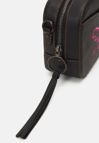 Černá kožená taška s hrubou texturou, bronzovým zipem, oranžovým prošíváním, detailem s třásněmi a růžovým logem blízko vršku.