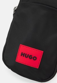 Schwarze Stofftasche mit einem roten rechteckigen Patch, auf dem das Wort "HUGO" in fetten schwarzen Buchstaben steht.