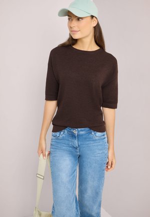 Kobieta w jasnoniebieskiej czapce, ciemnobrązowym swetrze z krótkim rękawem i wyblakłych niebieskich dżinsach, trzymająca jasny pasek torby, patrzy w prawo.