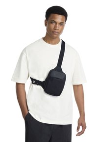 Black crossbody bag made of durable fabric, featuring a zip pocket, adjustable strap, and logo detail. Worn over a light-colored shirt.