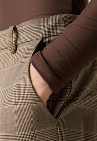 Un haut à manches longues marron est associé à un pantalon à carreaux avec un motif à check marron et beige. Le pantalon possède des passants de ceinture et des poches latérales.