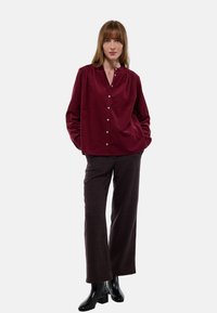 Chemise boutonnée bordeaux avec un col, détails froncés aux épaules, et une coupe décontractée, associée à un pantalon à carreaux sombres et des bottines noires.