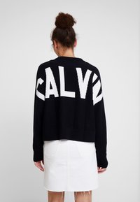 Czarny krótki sweter z dużym białym napisem "CALV" na plecach, wykonany z miękkiej dzianiny, zestawiony z białą spódnicą z denimu.