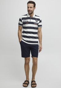 Camisa polo a rayas en azul marino y blanco, con cuello y detalles de emblema frontal, combinada con pantalones cortos azul marino y sandalias marrones.