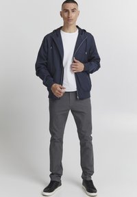 Jachetă cu glugă de culoare bleumarin, cu fermoar, purtată peste un tricou alb. Pantaloni gri și adidași negri completează ținuta. Textură netedă a țesăturii.