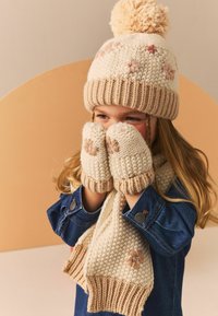 Strikket cremet vinterhat med pom-pom, matchende vanter og tørklæde. Har beige accenter og blomsterbroderi på vanterne og tørklædet.