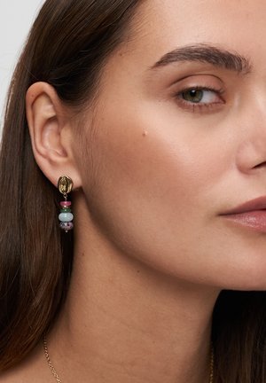 Nahaufnahme des Gesichts einer Frau, die einen goldenen Ohrring mit pinken, blauen und lila Perlen trägt, der von ihrem linken Ohr hängt.