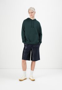 Fiatal személy rövid szőke hajjal, sötétzöld kapucnis pulóvert, sötétkék fényes térdnadrágot, fehér zoknit és krémszínű sportcipőt visel.