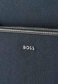 Κοντινή λήψη σκούρου υφάσματος με υφή, με ασημί φερμουάρ και μικρά μεταλλικά γράμματα που σχηματίζουν τη λέξη «BOSS» στην επιφάνεια.