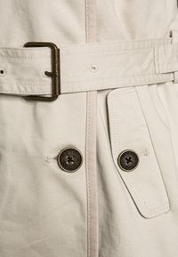 Detalle de un abrigo trench beige que muestra una tela resistente con una textura suave, con una hebilla de metal brillante y dos botones redondos grandes.