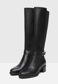 Bottes en cuir noir arrivant jusqu'au genou avec une texture lisse, bout pointu, talon bloc, et une sangle sur le côté dotée d'un accent métallique.