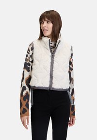 Weißes gesteppte Weste mit hohem Kragen, weichem Stoff und grauen Besätzen, kombiniert mit einem gemusterten Langarmoberteil mit braunen und schwarzen Leopardenmotiven.